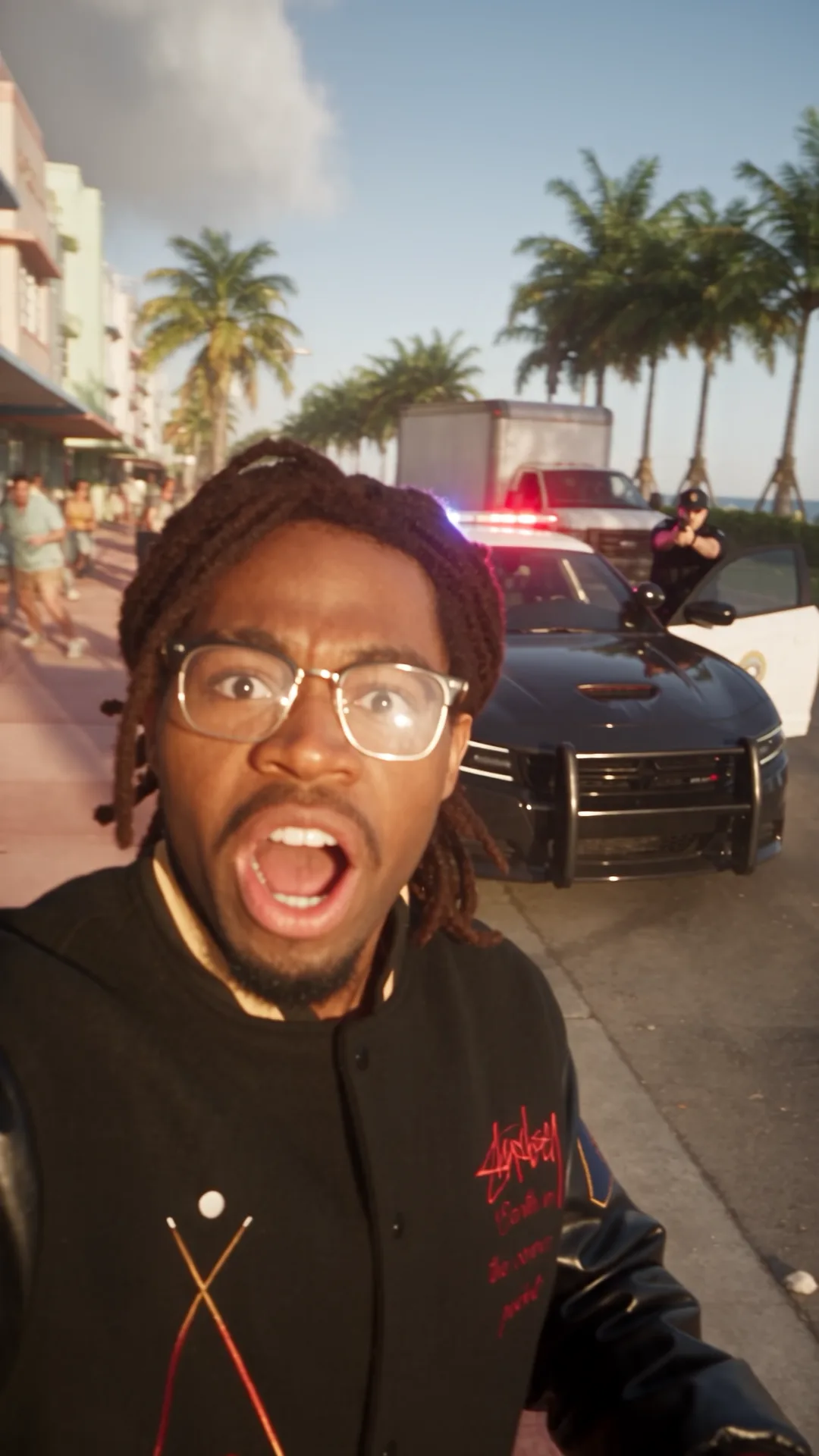 A man in a black varsity jacket films himself in shock on a palm-lined Art Deco boulevard as a police officer aims a weapon beside a cruiser