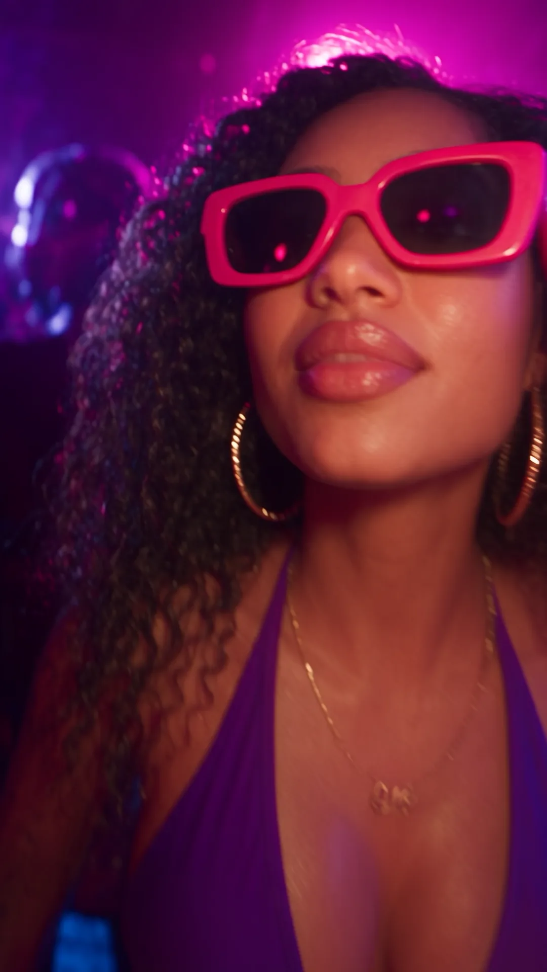 A woman in pink sunglasses and a purple top lit by magenta club lighting, a mirror ball glowing behind her