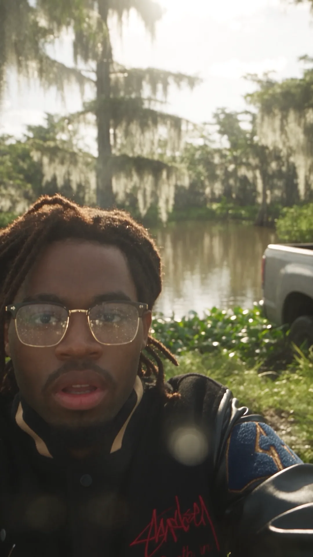 The same man films himself beside still swamp water under cypress trees draped in Spanish moss, a pickup truck parked behind him